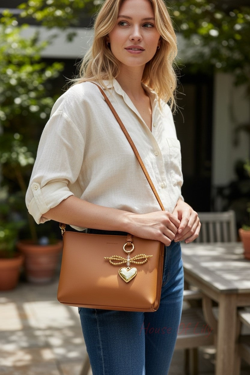 A golden keychain featuring a beaded bow and heart charm hanging from a tan leather shoulder bag.