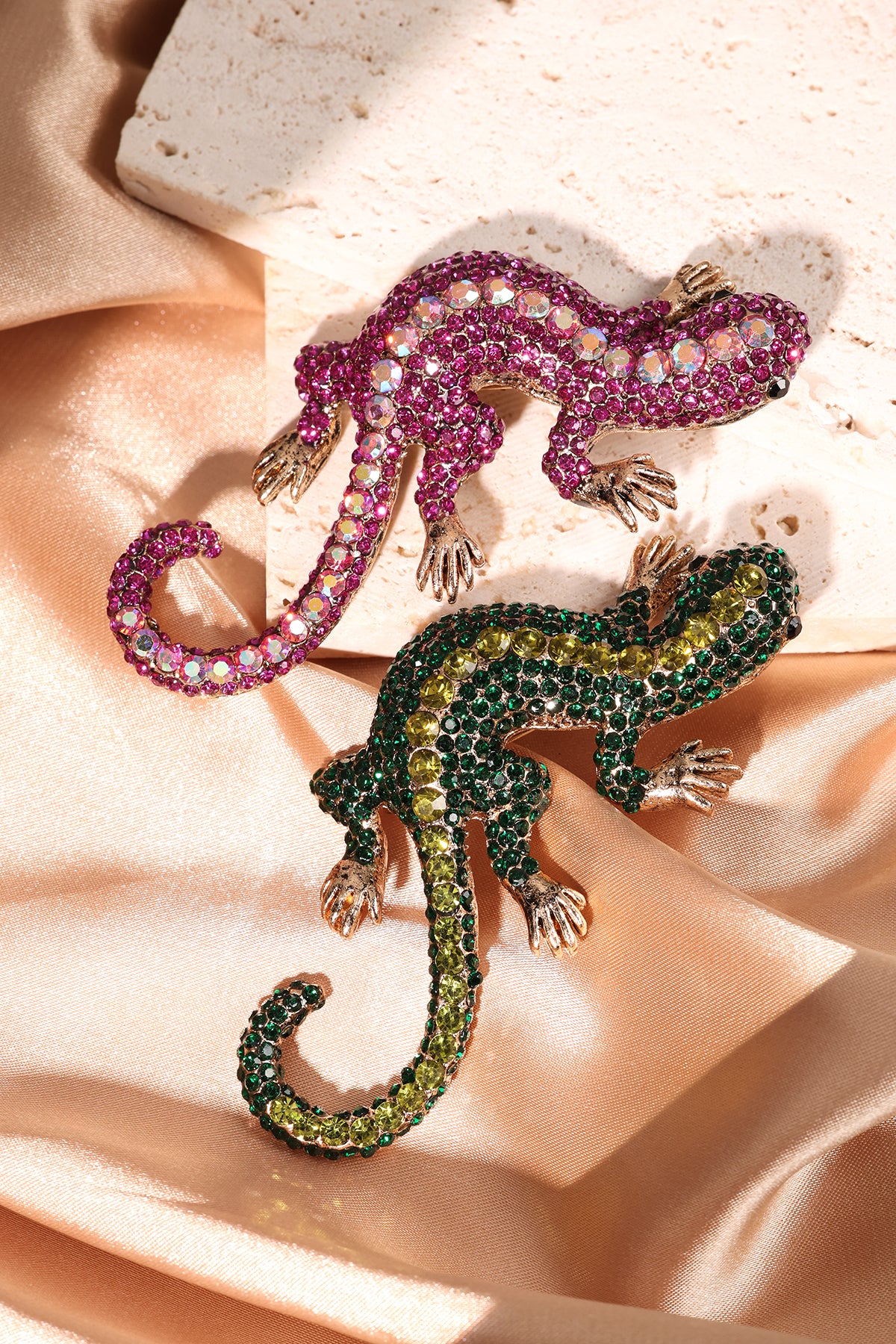 Two gecko brooches, green with yellow crystals and fuchsia with iridescent crystals, on silky fabric and stone