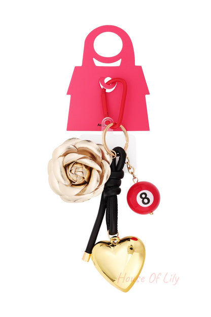 Product layout view of the bag charm hanging from red packaging card, displaying the gold rose, red 8-ball, glossy golden heart, and red clip