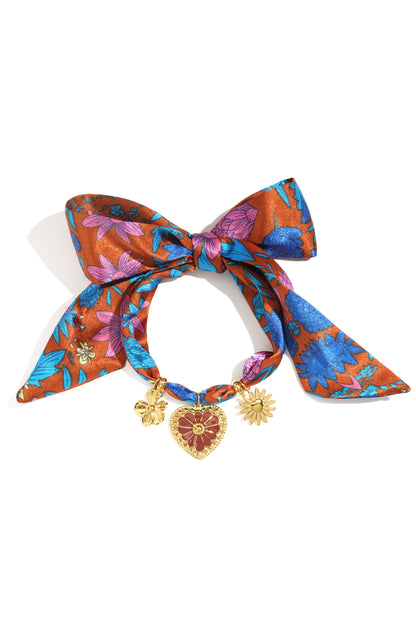Close-up of a floral charm bracelet featuring a burnt orange fabric with blue and pink flowers tied in a bow, adorned with gold-tone flower and heart charms