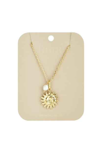 Gold sun face pendant necklace on a chain with a small white baroque pearl charm on a branded beige display card.