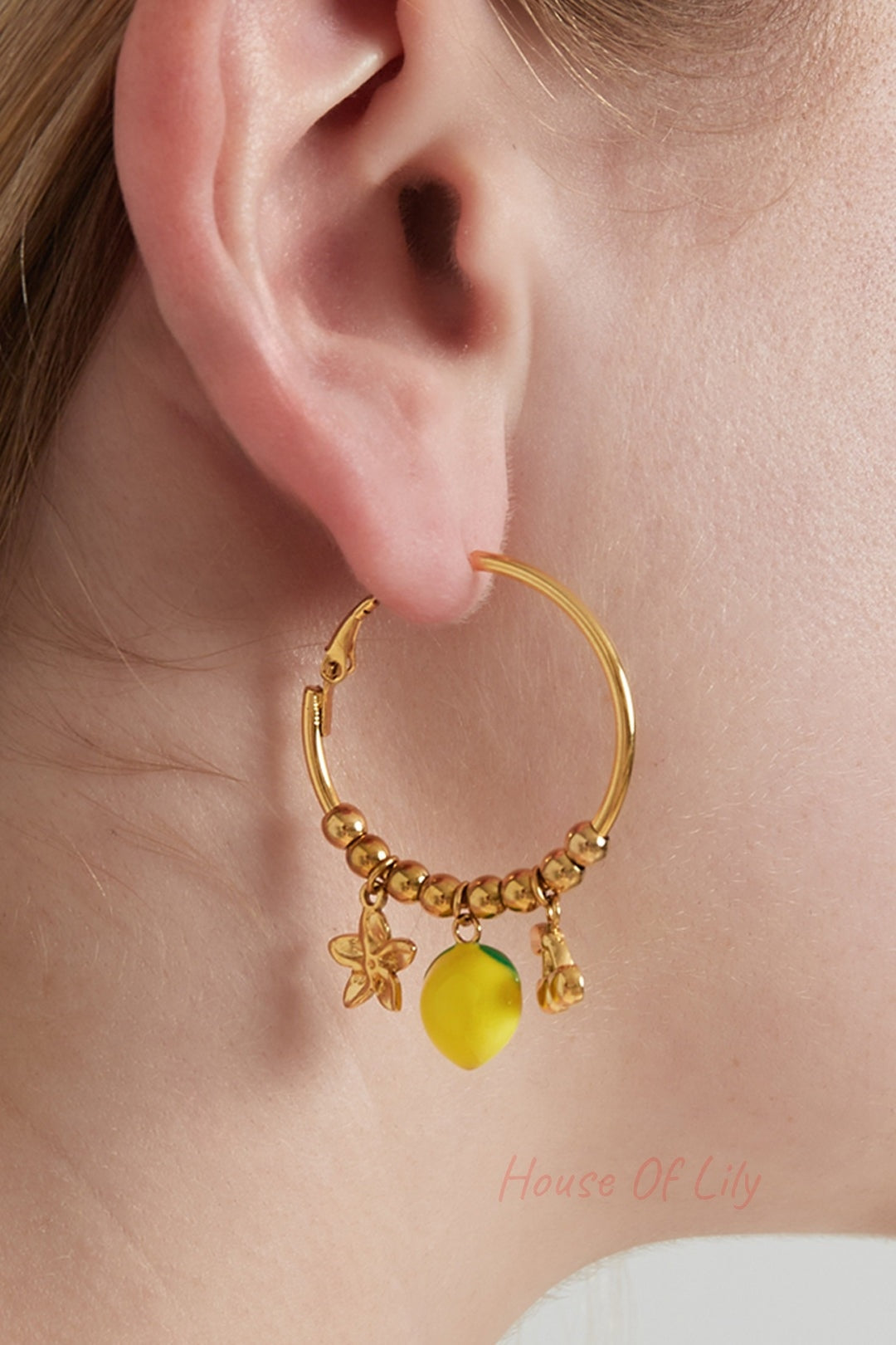 Close-up of a woman wearing gold-tone hoop earring with playful fruit charms including a yellow lemon, gold cherries, and a flower charm