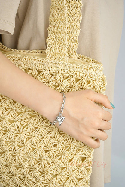 Silver chunky heart charm bracelet worn on wrist holding a woven beige tote bag