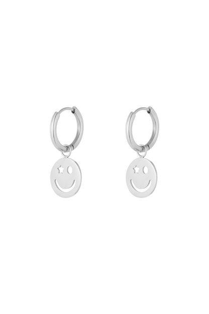 Pair of silver hoop earrings with dangling smiley face charm featuring one star eye and one circular eye on white background