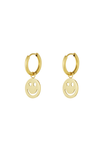 Pair of gold hoop earrings with dangling smiley face charm featuring one star eye and one circular eye on white background