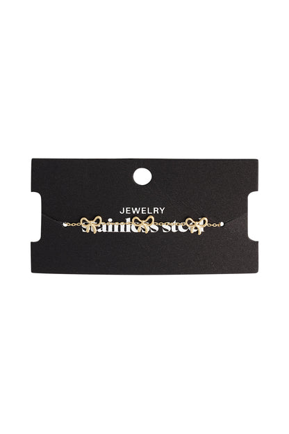 Gold bow charm bracelet displayed on black jewelry card with 'stainless steel' branding