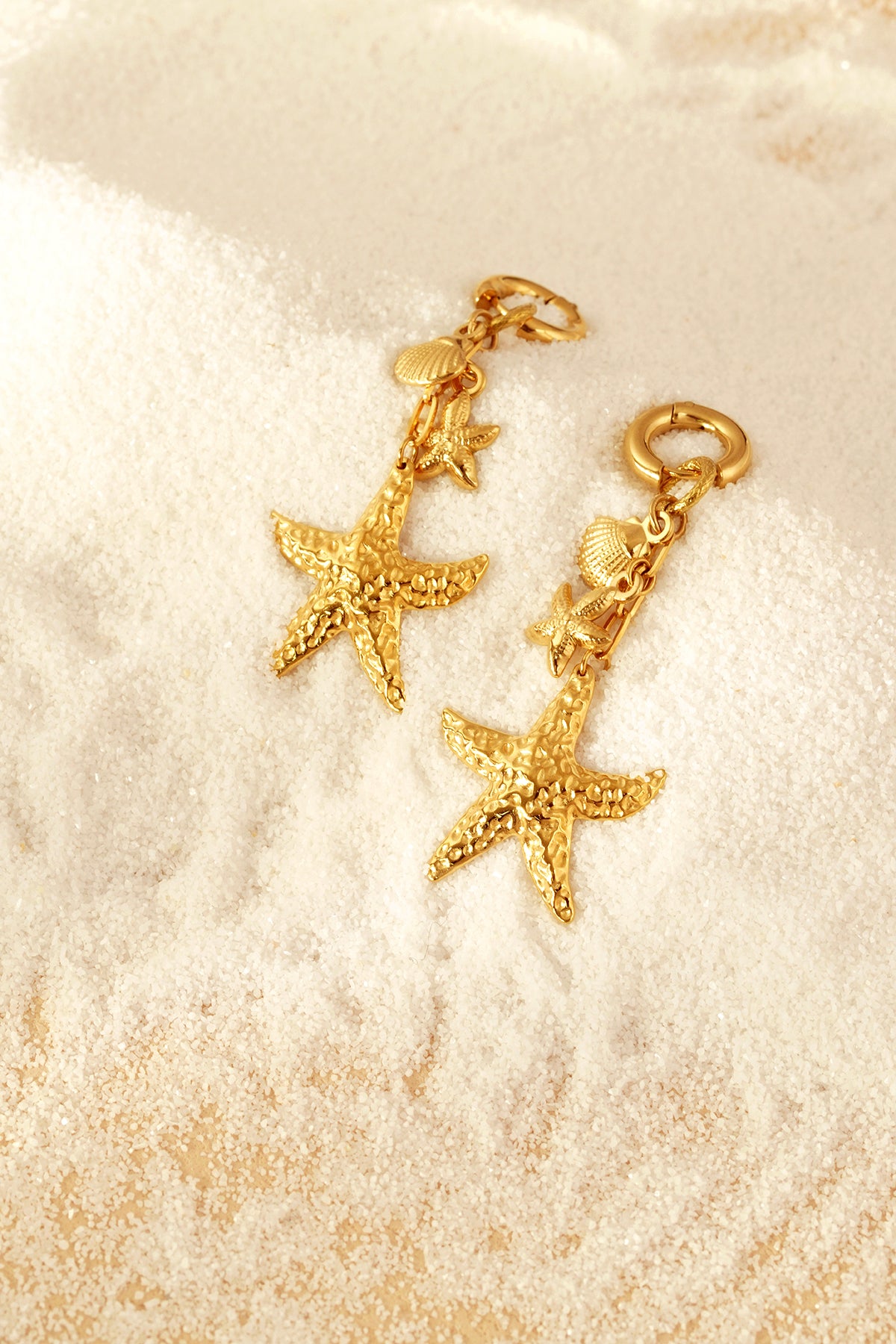 Gold starfish and seashell drop earrings resting on fine white sand