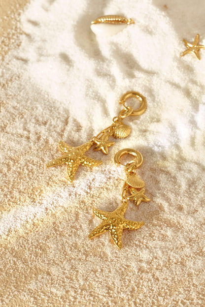 Close top-angle view of gold starfish and seashell drop earrings on textured pale sand
