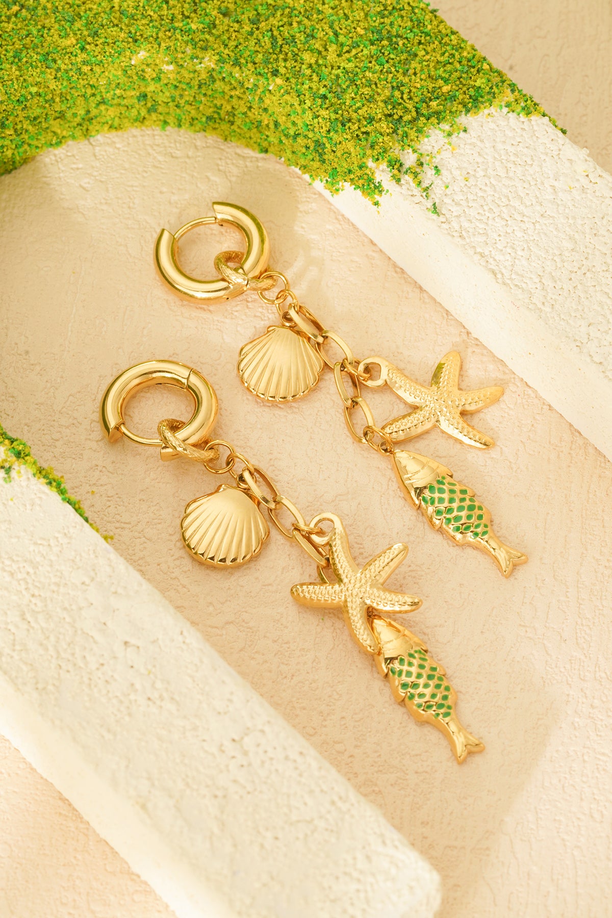Pair of gold-tone sea charm drop earrings with starfish, seashell and green enamel fish charms styled on beige textured surface with green moss