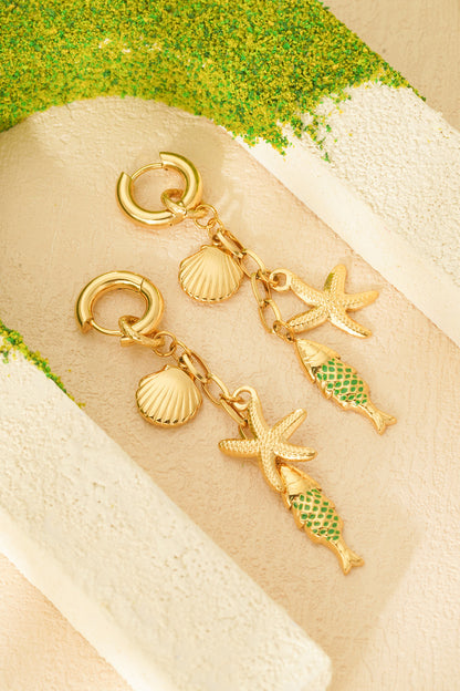 Pair of gold-tone sea charm drop earrings with starfish, seashell and green enamel fish charms styled on beige textured surface with green moss