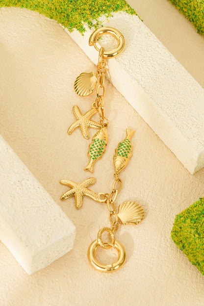 Gold-tone sea charm bracelet with starfish, seashell and green enamel fish charms styled on textured beige surface with green moss accents