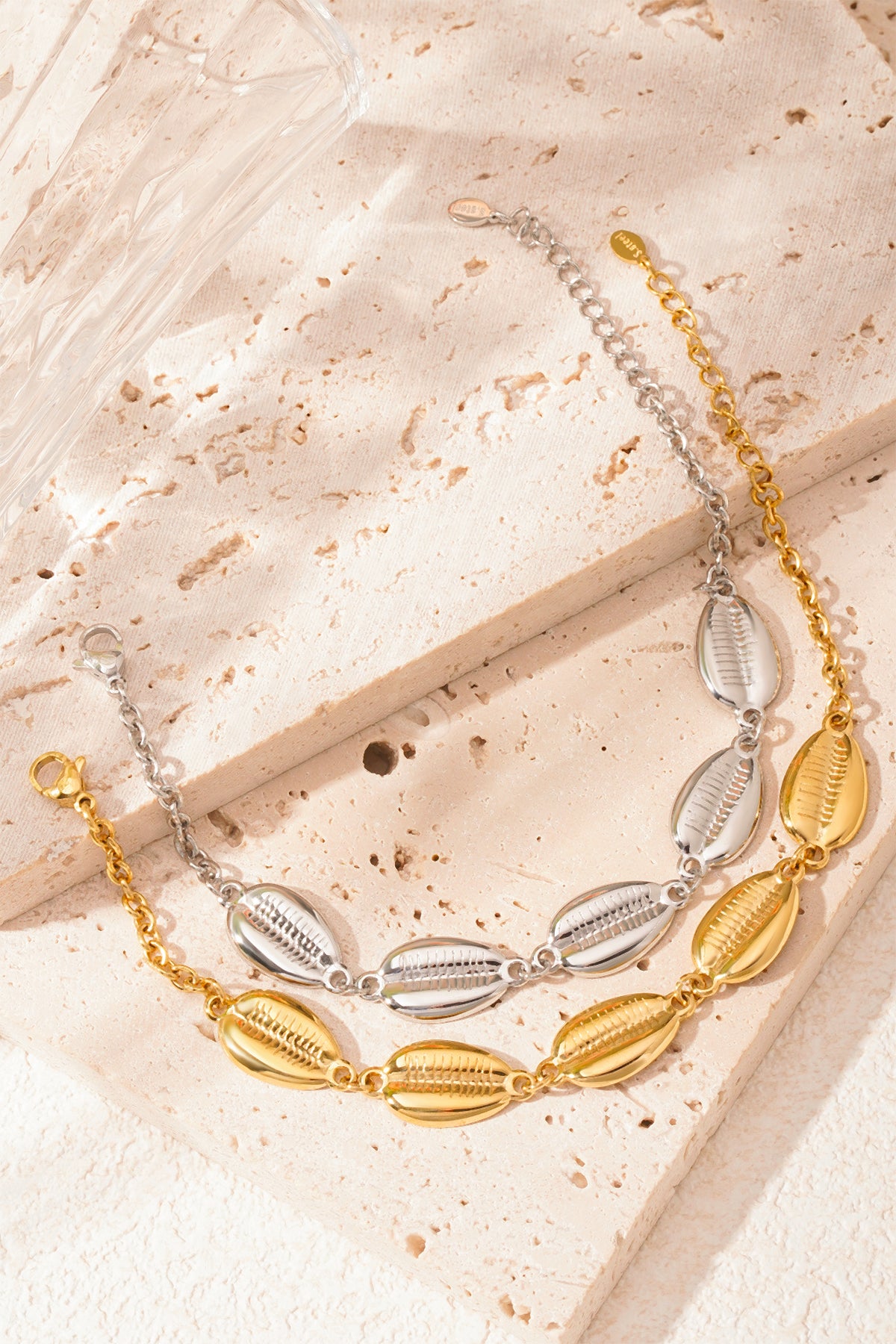 Silver-tone and gold-tone shell charm bracelets laid flat on a rustic stone surface with soft sunlight illumination