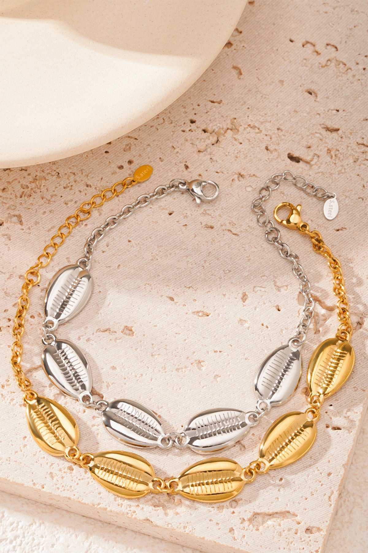 Two shell charm bracelets displayed side by side, one in silver-tone and one in gold-tone, on a textured beige surface