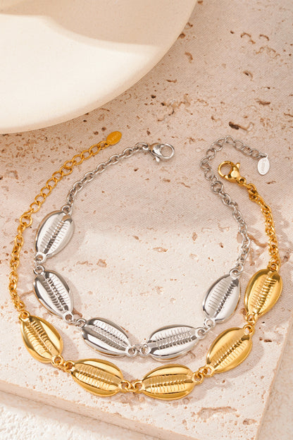 Two shell charm bracelets displayed side by side, one in silver-tone and one in gold-tone, on a textured beige surface