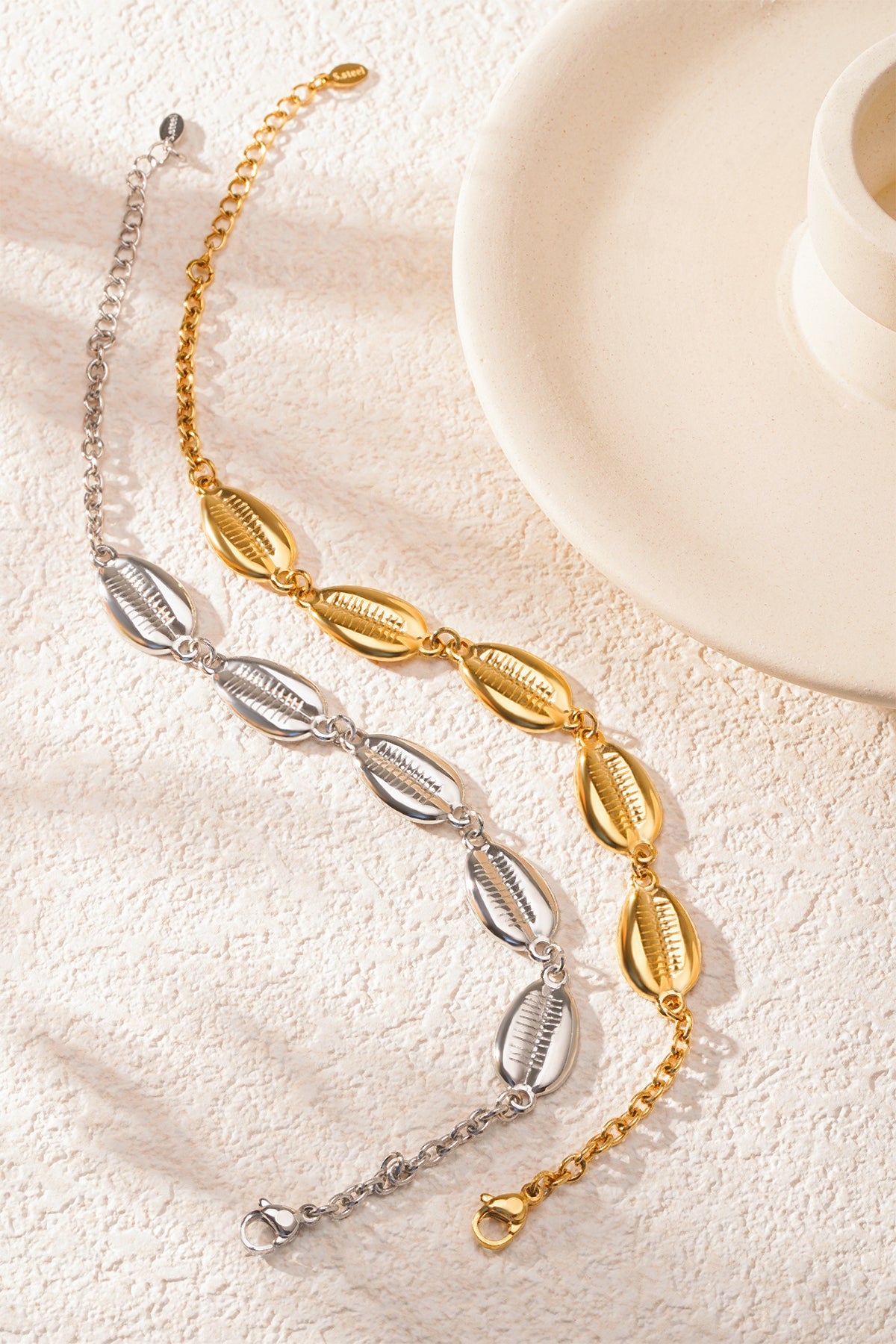 Golden and silver shell charm bracelets elegantly arranged beside a cream ceramic bowl on a light textured background