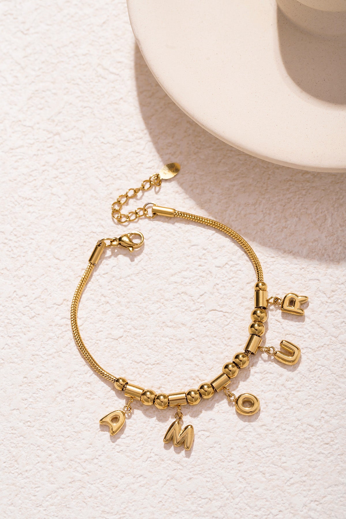 Detailed close-up of gold-tone AMOUR charm bracelet showing polished beads and letter charms on light stone background