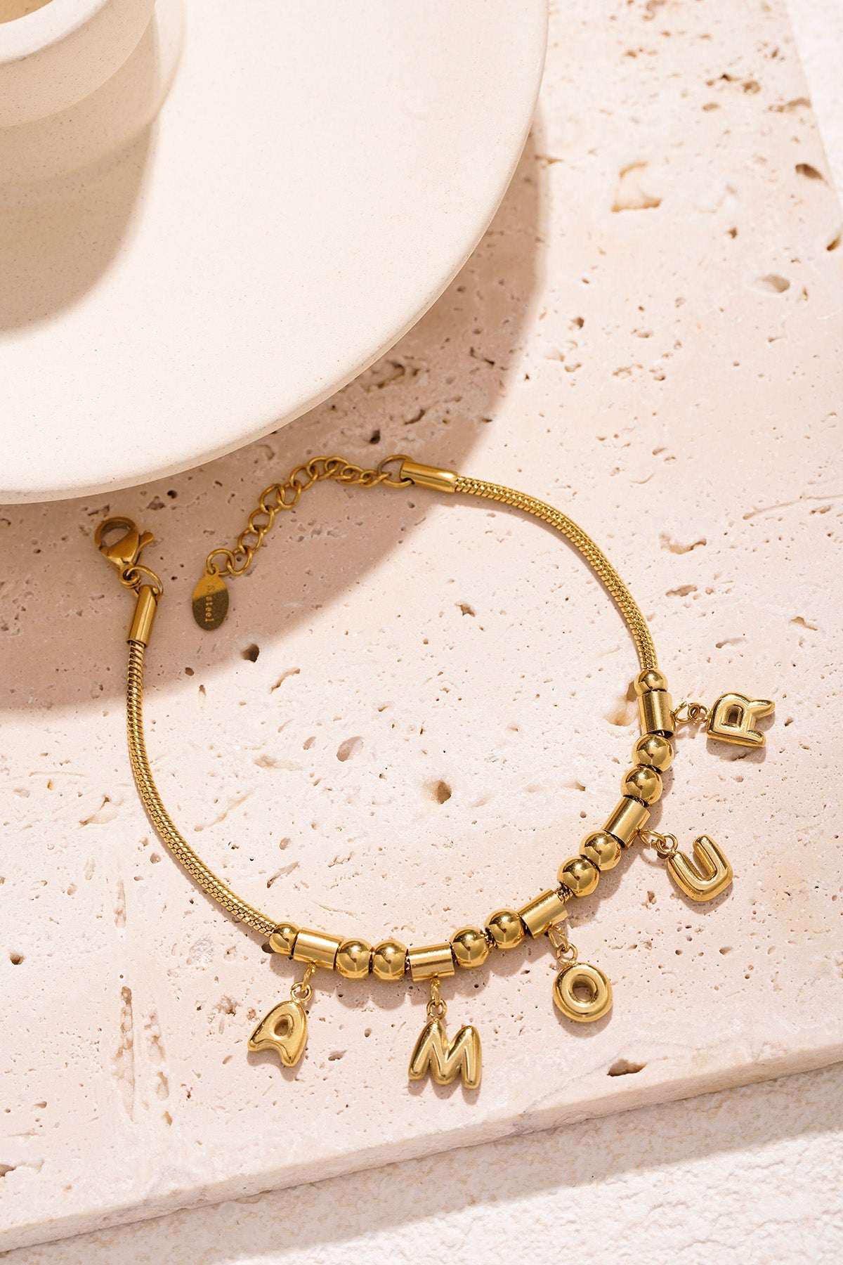 Gold-tone AMOUR charm bracelet styled on a textured light stone surface next to a white cup and saucer