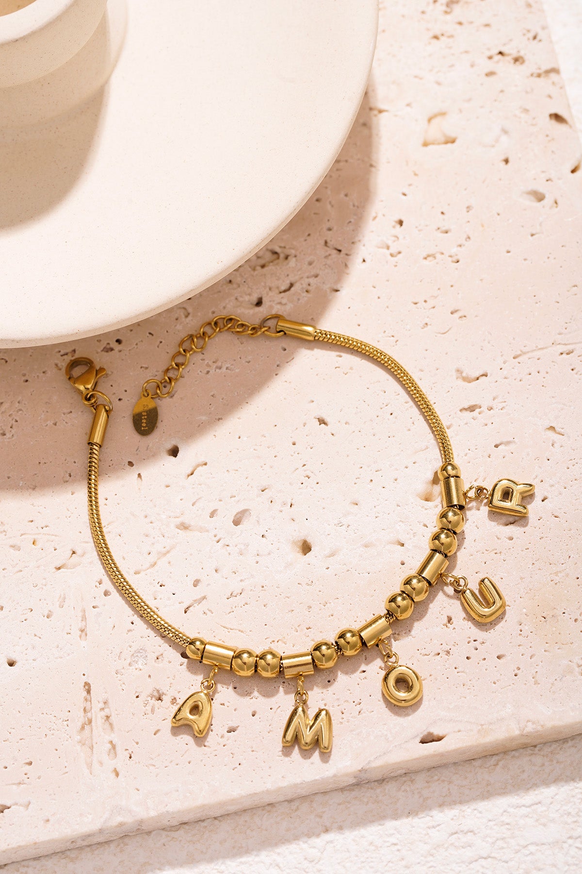 Gold-tone AMOUR charm bracelet styled on a textured light stone surface next to a white cup and saucer