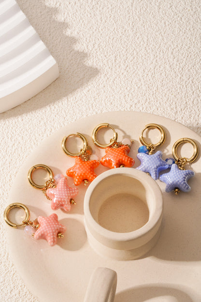 Array of starfish charm hoop earrings in coral pink, baby blue, and bright orange arranged beside a cream ceramic holder