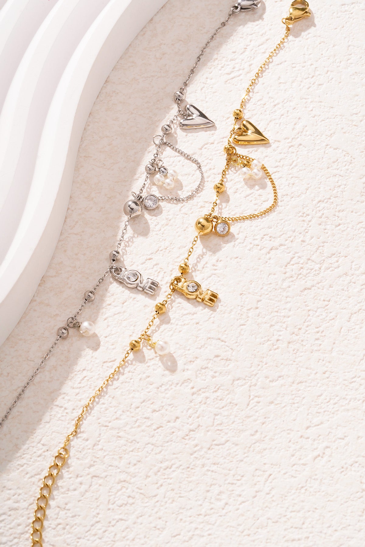 Silver and gold tone charm bracelets lying side by side on a creamy textured surface highlighting the heart, pearl, and LOVE charms