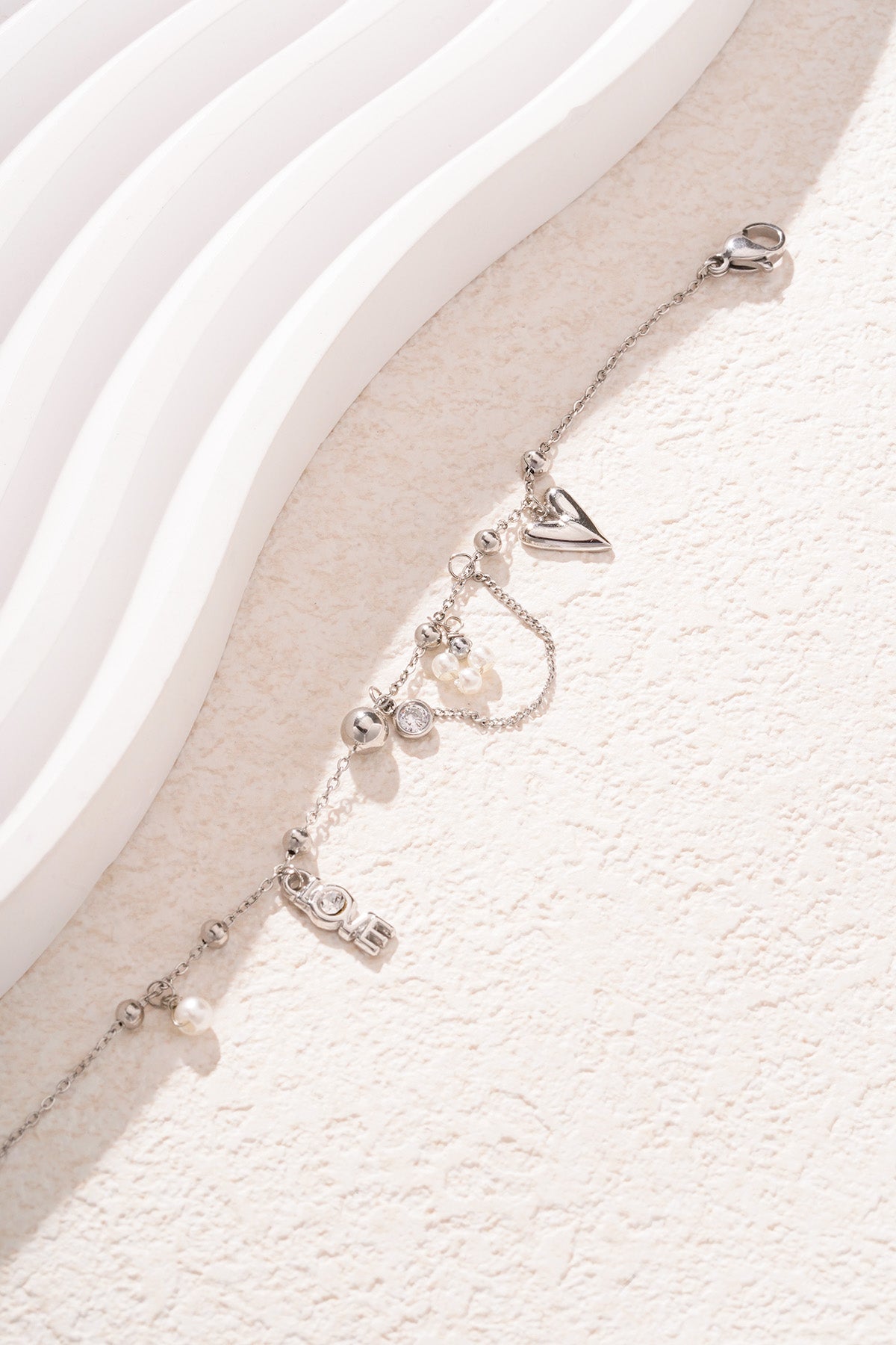 Close up of silver-tone charm bracelet featuring heart, LOVE letter crystals, and faux pearl accents on a light textured background