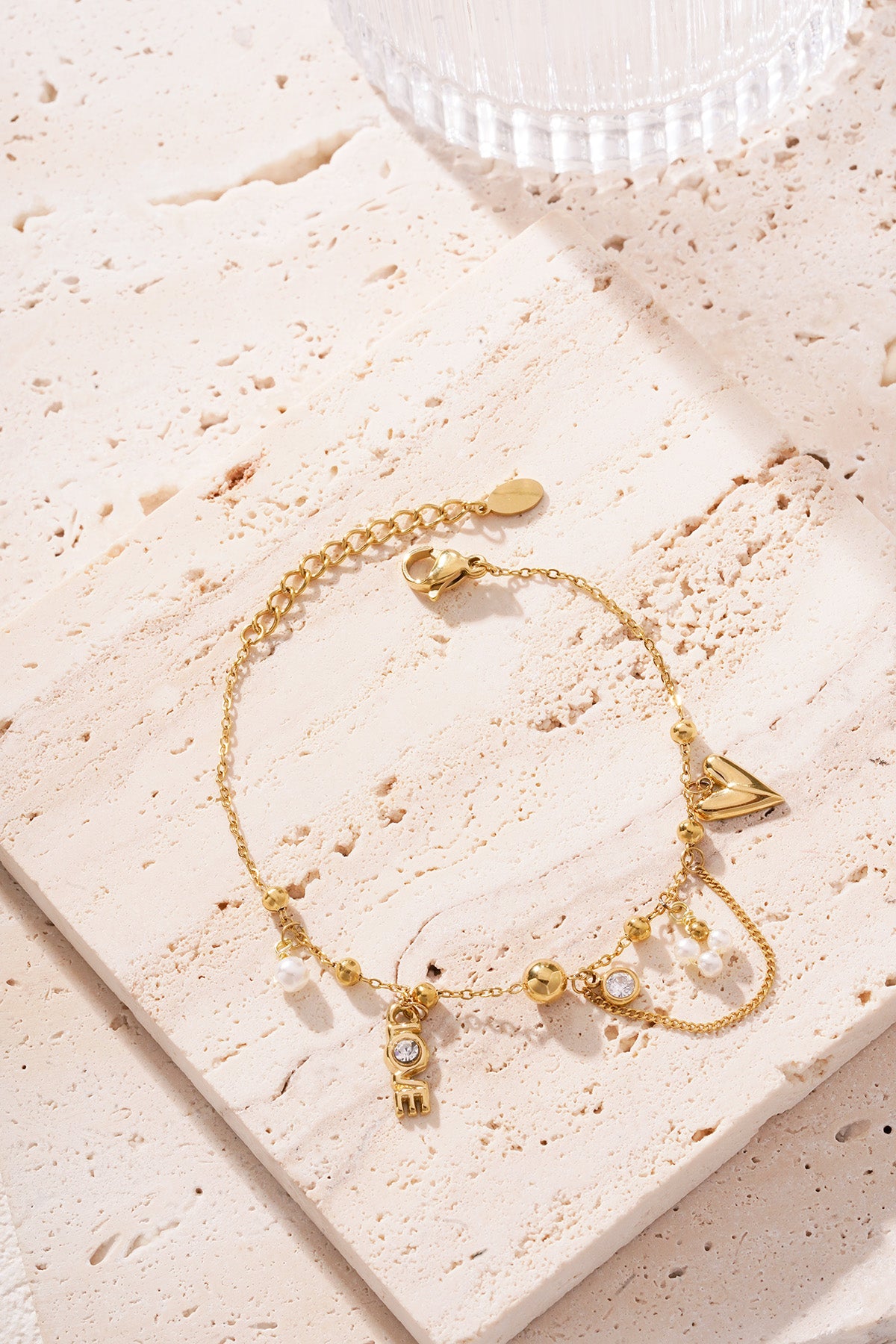 Close shot of gold-tone charm bracelet on textured beige fabric next to a glass