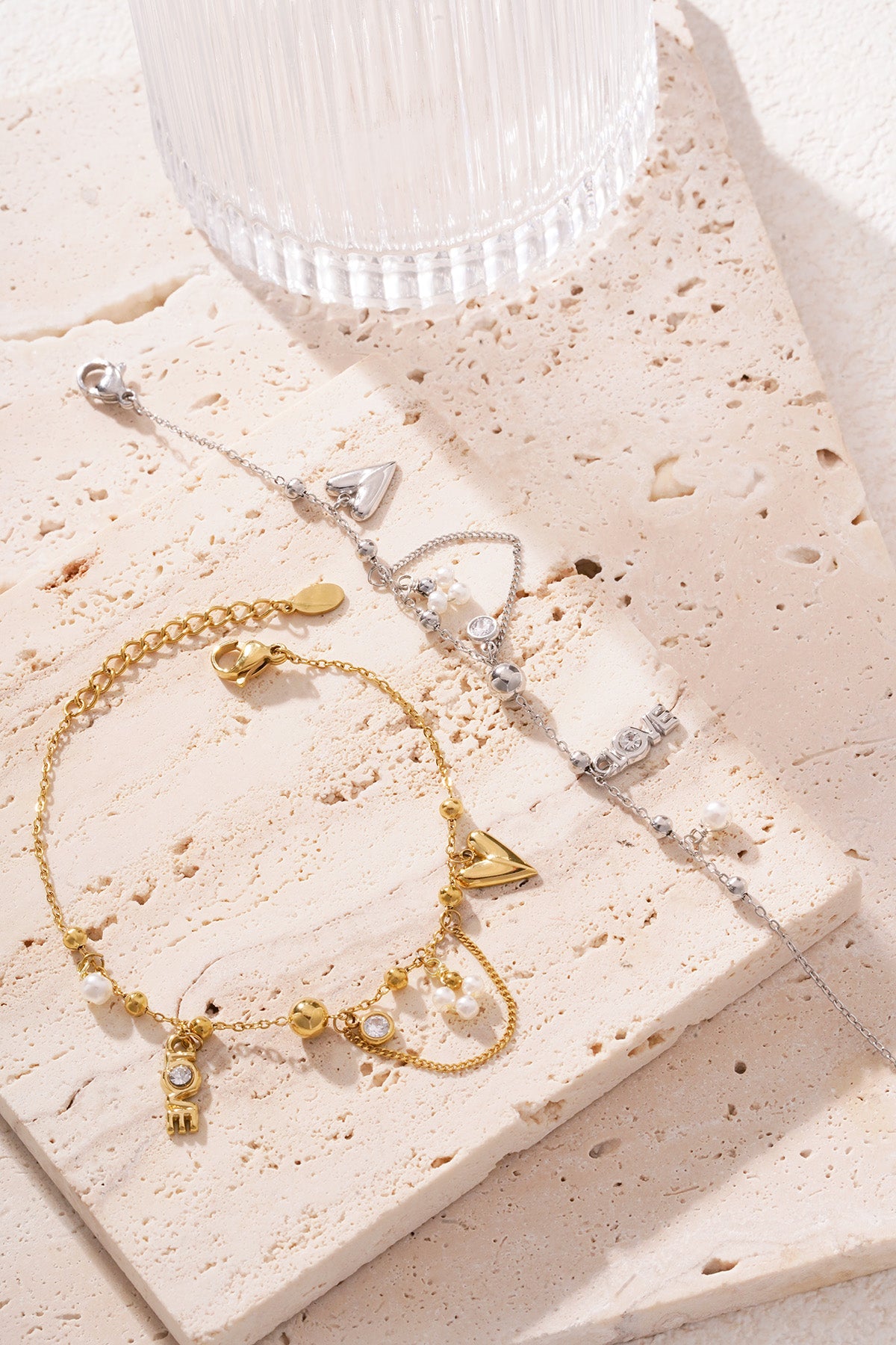 Silver-tone charm bracelet lying beside a gold-tone one on a cream textured stone surface with sunlight highlighting the details
