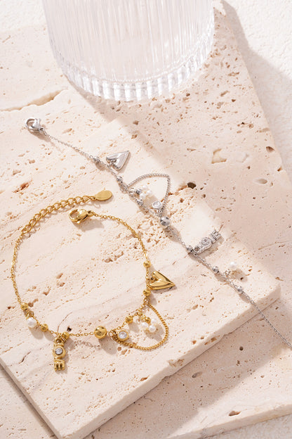 Silver-tone charm bracelet lying beside a gold-tone one on a cream textured stone surface with sunlight highlighting the details