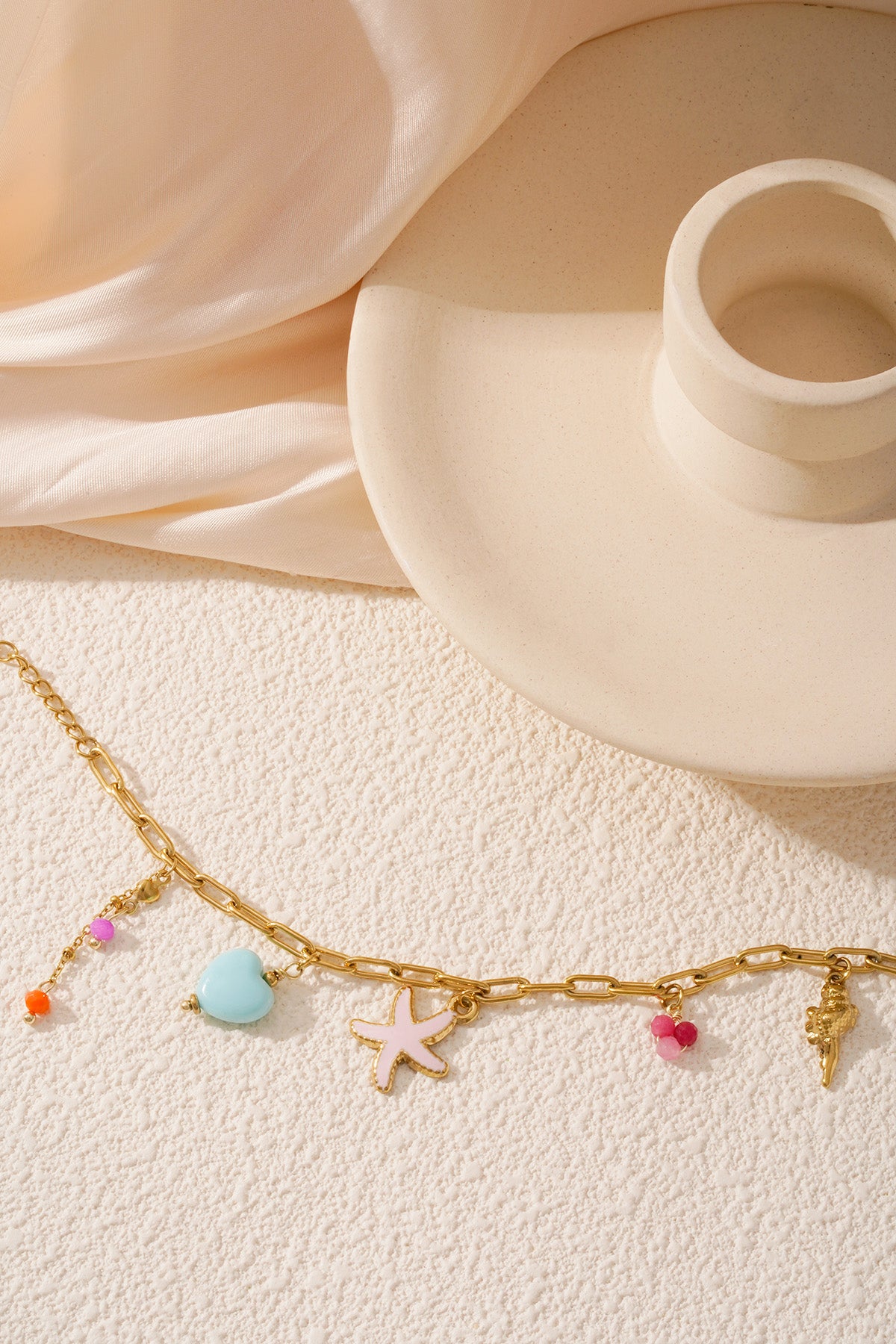 Gold charm bracelet laid on textured surface near cream-coloured candle holder with starfish, heart, bead, and seashell charms