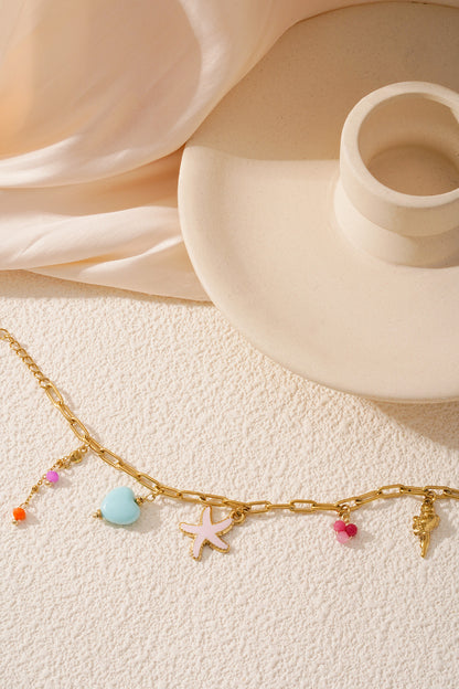 Gold charm bracelet laid on textured surface near cream-coloured candle holder with starfish, heart, bead, and seashell charms