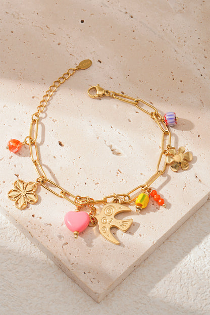 Golden link bracelet with colourful beads and flower, bird and heart charms laid on textured cream surface