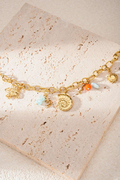Bracelet laid on textured cream stone tile highlighting gold chain, shell and sun charms with colourful beads