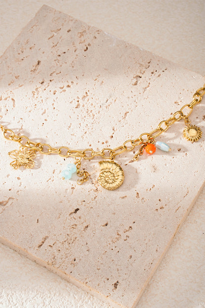 Bracelet laid on textured cream stone tile highlighting gold chain, shell and sun charms with colourful beads