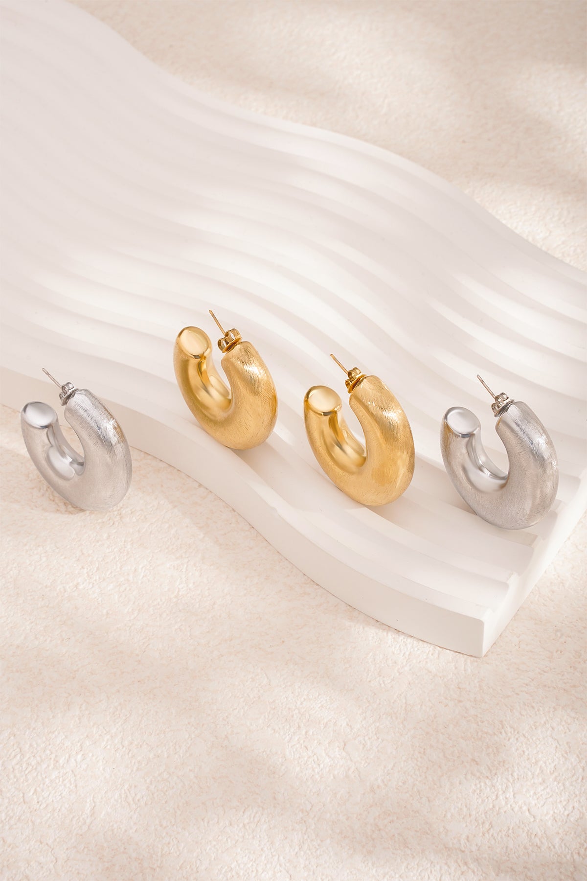 Close-up of two pairs of chunky hoop earrings, gold and silver, on sculptural white display