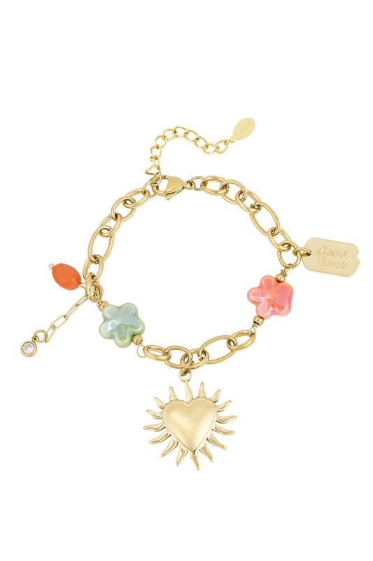 Gold-tone charm bracelet with heart sunburst pendant, green and coral flower-shaped beads, orange accent bead, and 'Good Luck' tag on white background