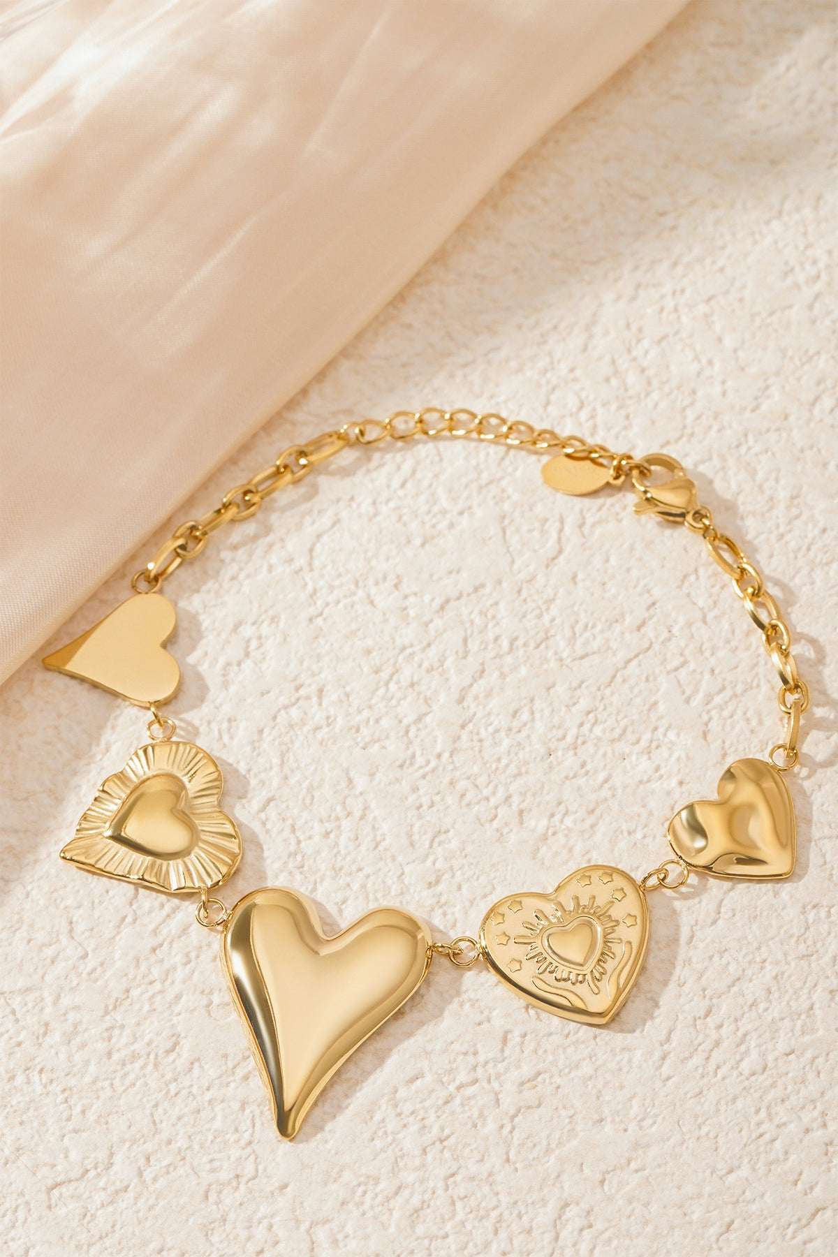 Gold chunky heart charm bracelet styled on a textured cream surface next to soft fabric, highlighting the shiny heart charms