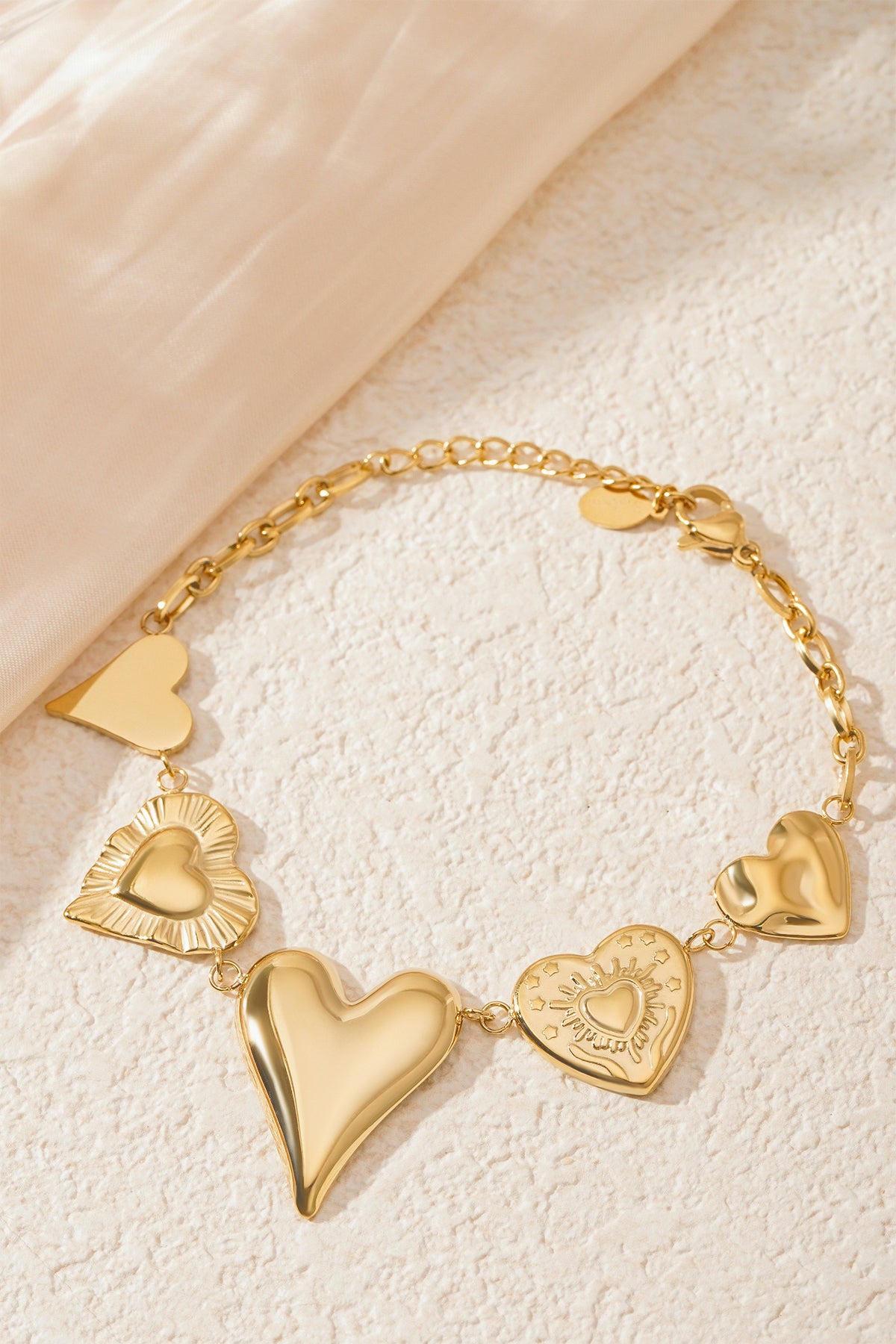 Gold chunky heart charm bracelet styled on a textured cream surface next to soft fabric, highlighting the shiny heart charms