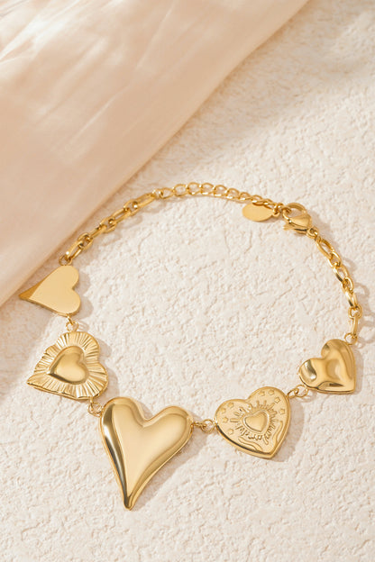 Gold chunky heart charm bracelet styled on a textured cream surface next to soft fabric, highlighting the shiny heart charms
