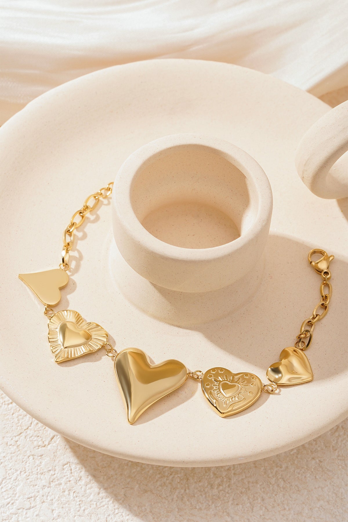 Gold chunky heart charm bracelet elegantly arranged on a cream decorative plate showing heart charms and lobster clasp