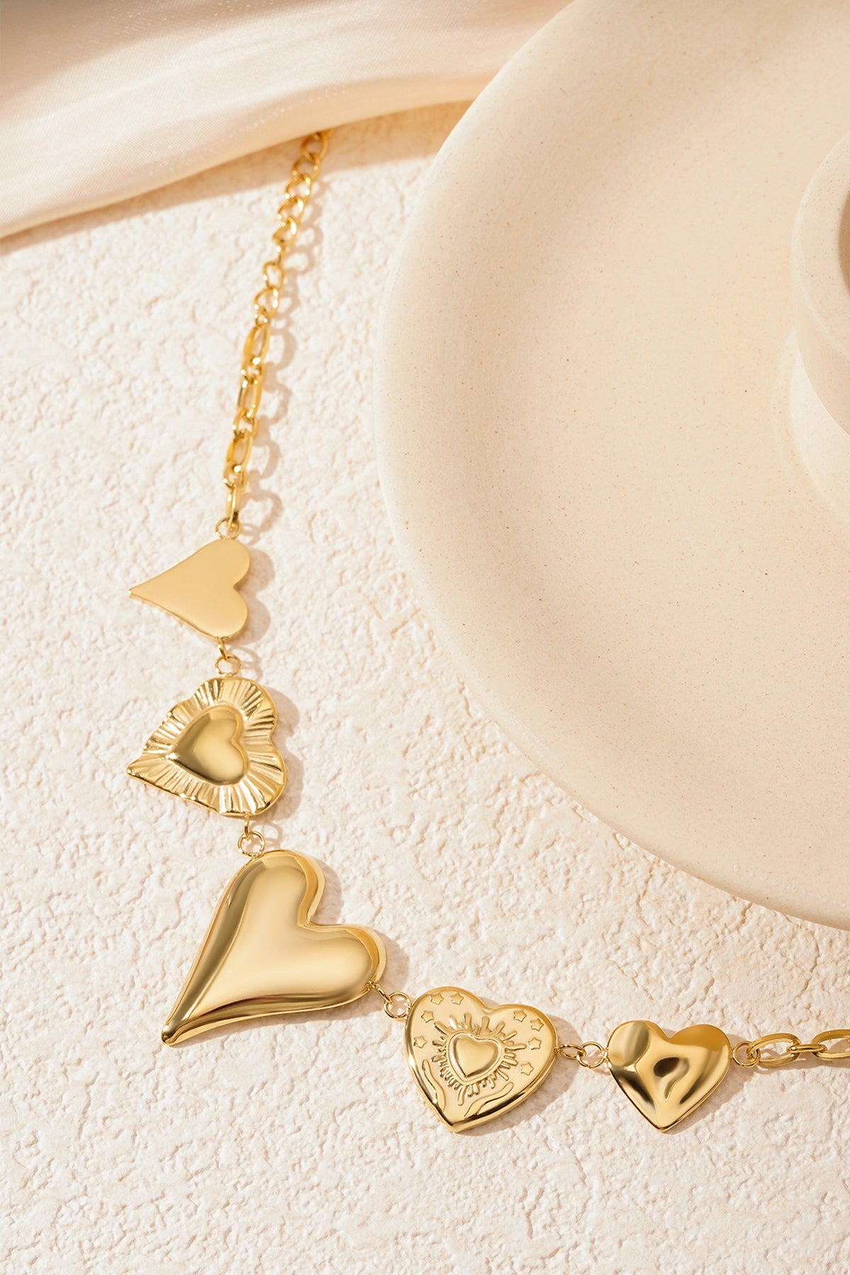 Gold chunky heart charm bracelet close-up on a cream textured surface beside a circular white decorative object