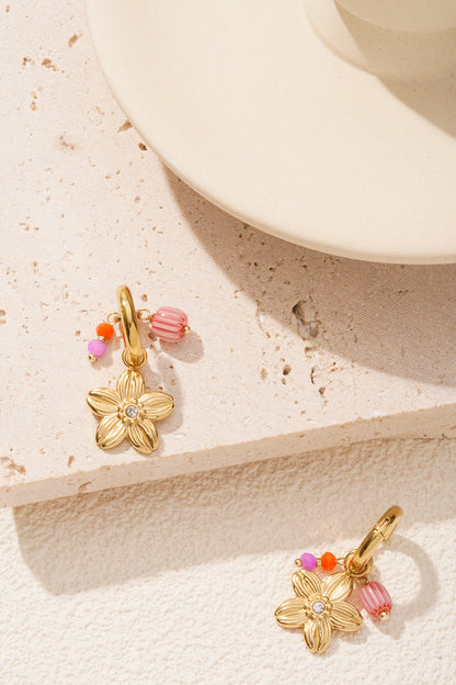 Gold-tone floral charm hoop earrings with colourful beads styled on a textured light stone surface near a cream ceramic object