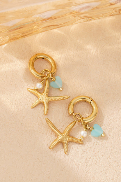 Gold hoop earrings with starfish charms, pearl bead, and aqua heart charm resting on textured beige surface