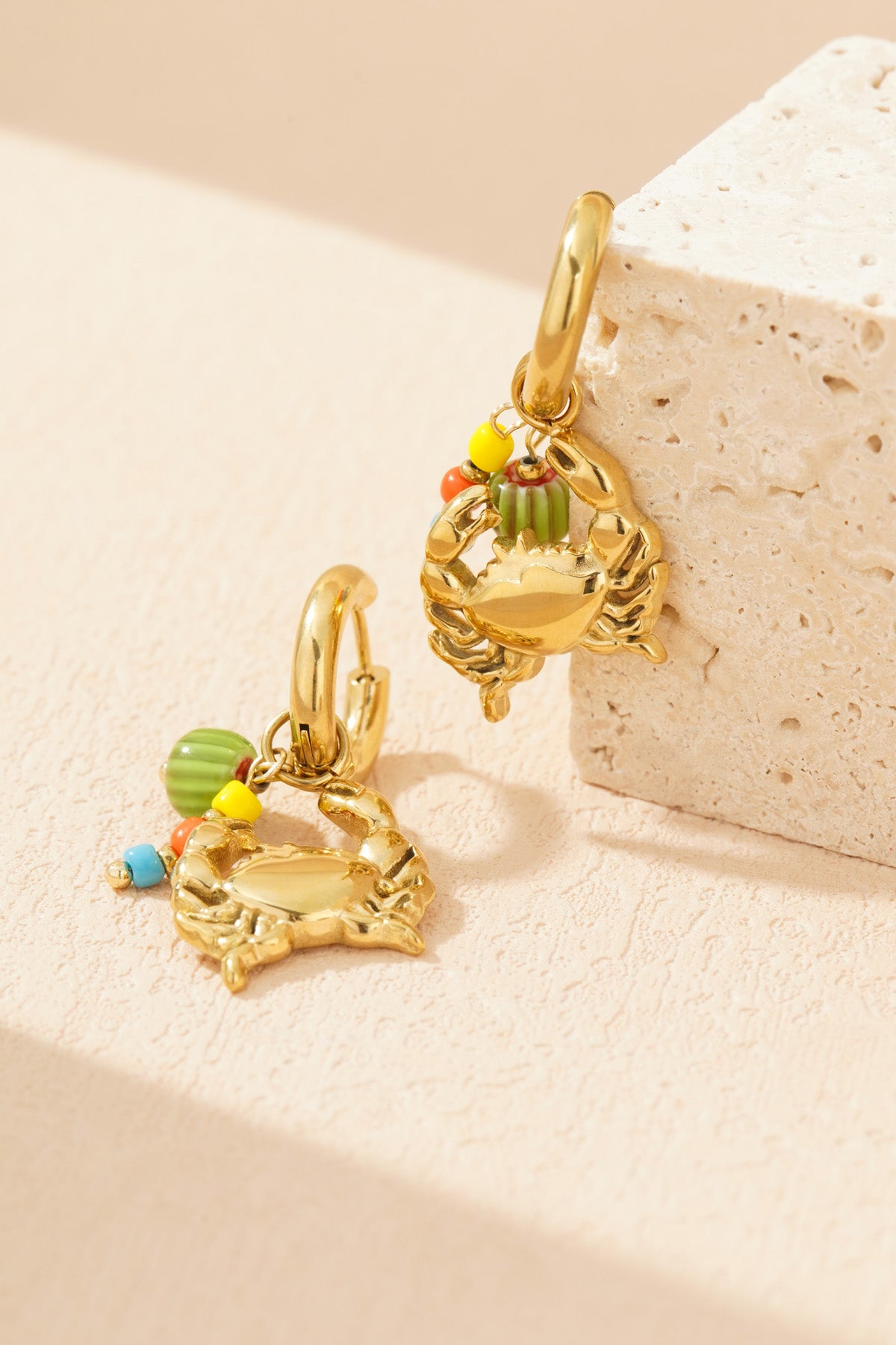 Gold-tone crab charm earrings with colourful beads resting against a textured stone surface