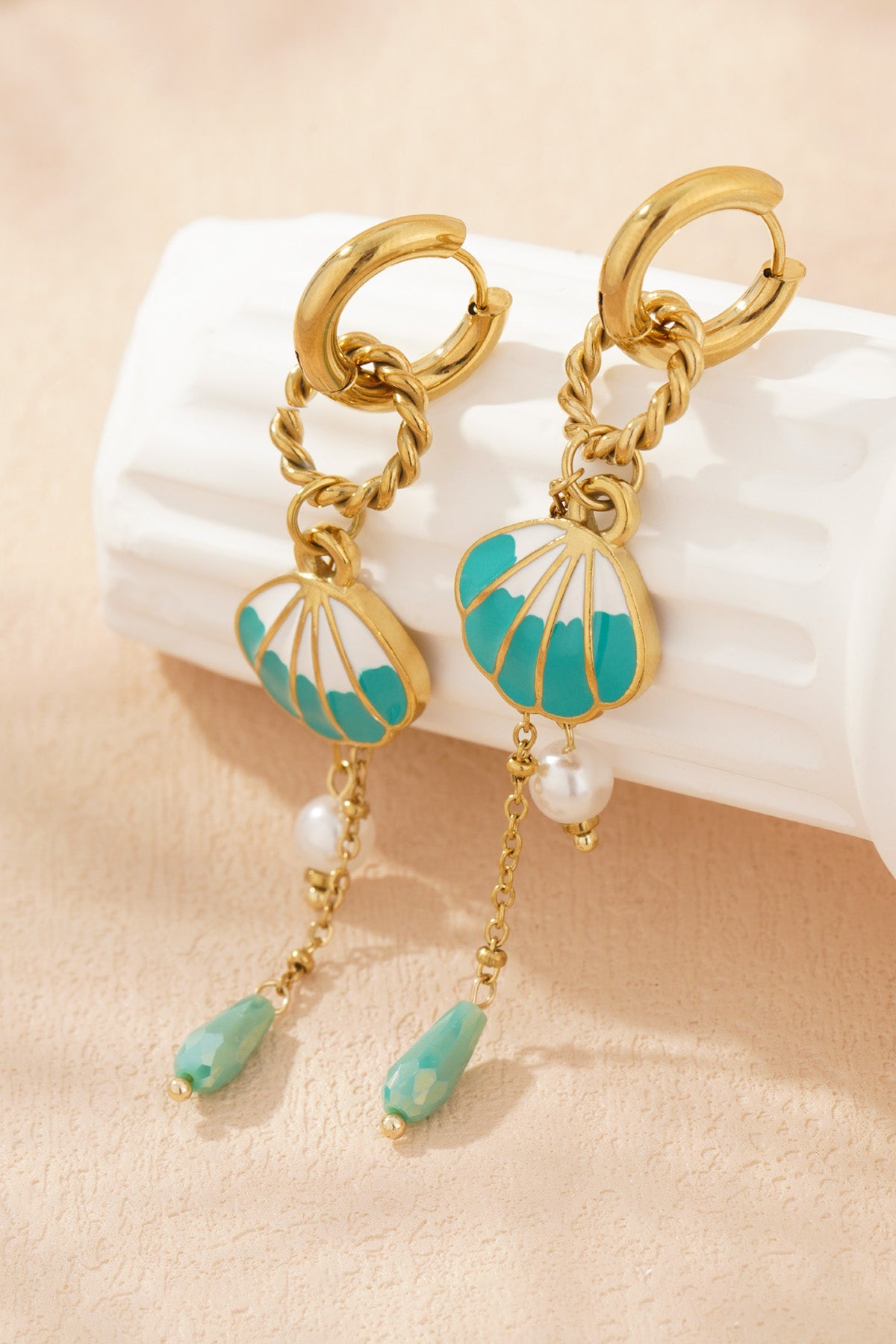 Shell charm drop earrings resting on textured cream background highlighting gold-tone shine, turquoise enamel shell and pearl bead details