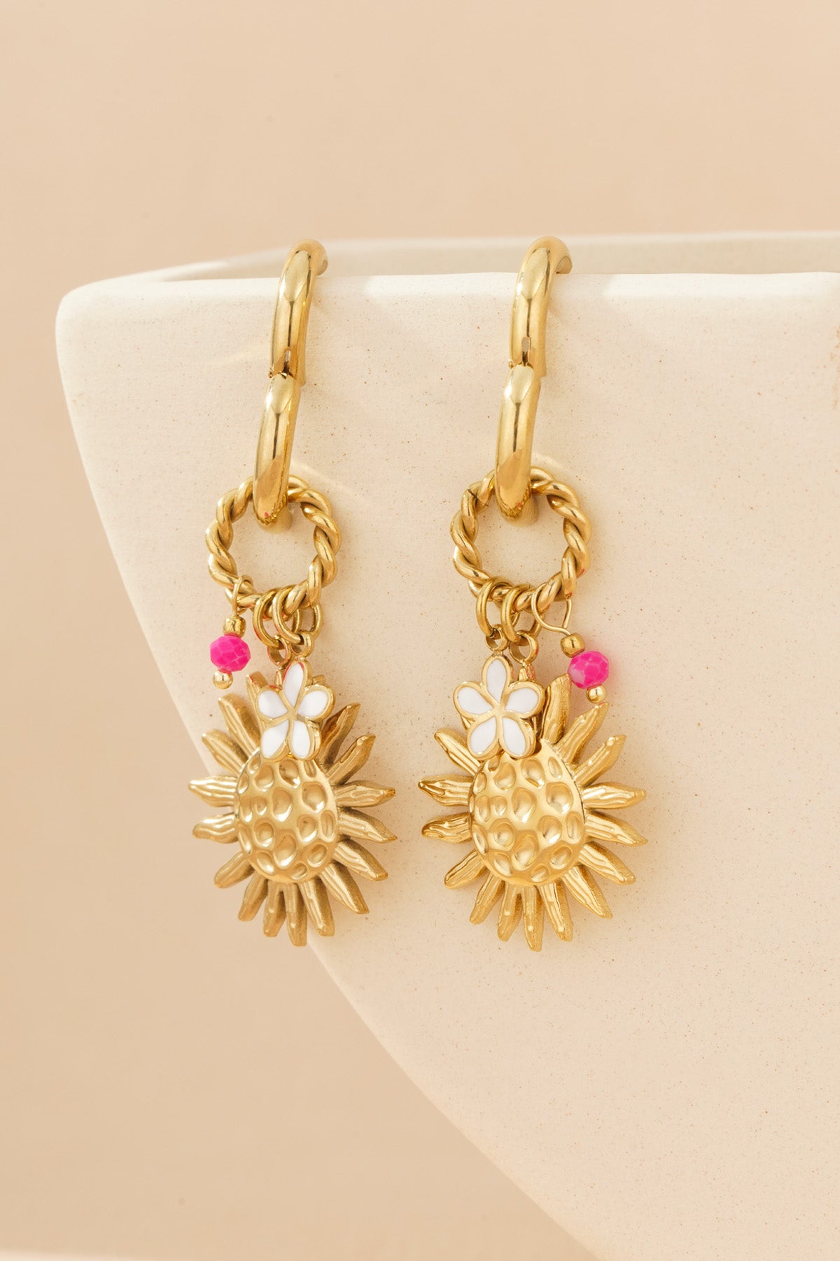 Gold-tone sunshine charm drop earrings hanging on cream-coloured pottery, showing textured sun designs with enamel flowers and pink beads.