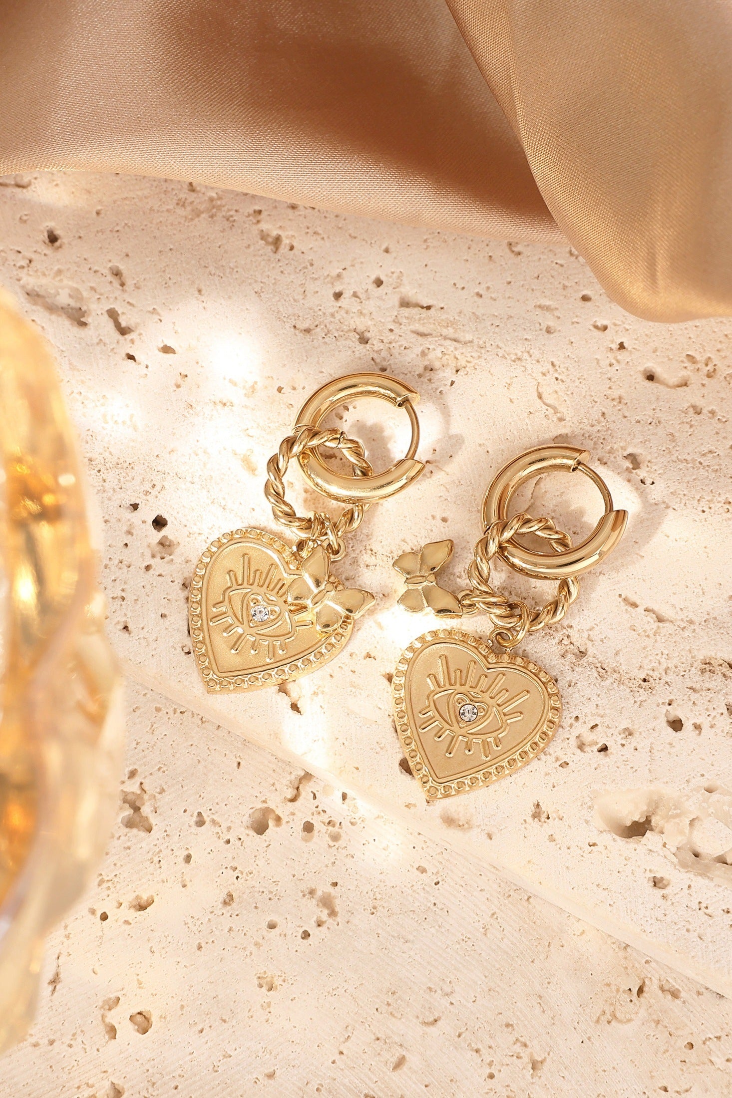 Gold heart charm hoop earrings lying on textured cream stone surface with sunlight