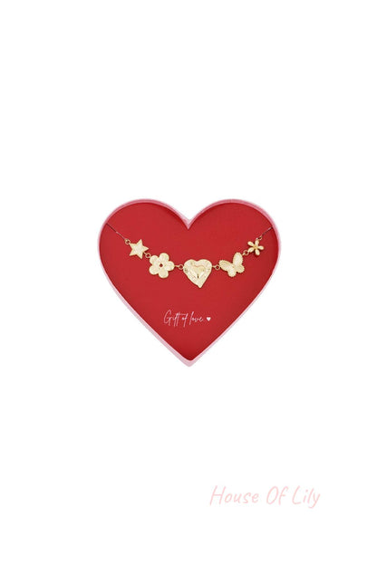 Gold charm bracelet with star, flower, heart, butterfly, and flower charms displayed in a red heart-shaped box with 'Gift of love' text