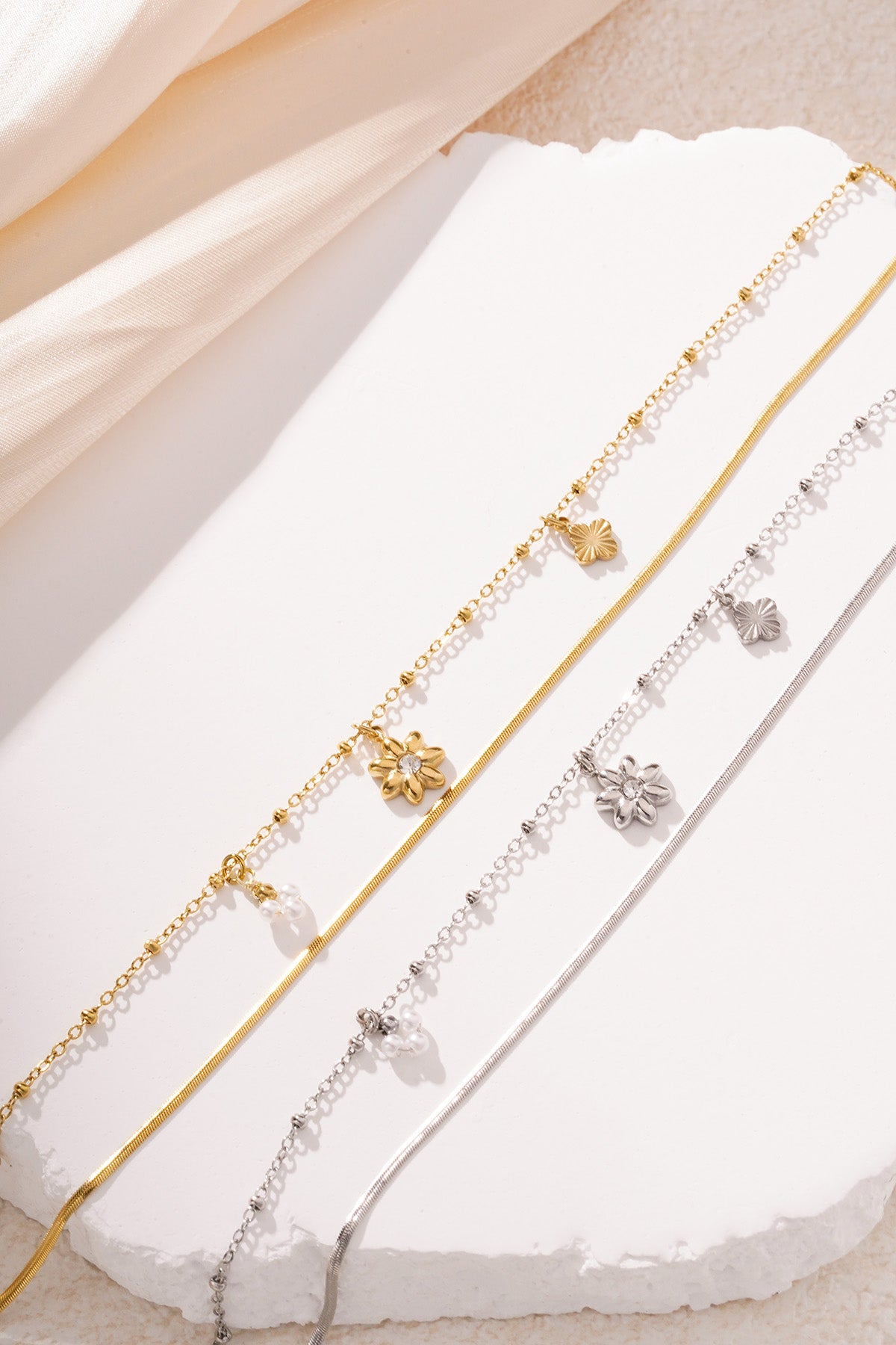 Gold and silver versions of layered floral charm bracelets with pearl and flower charms displayed on white textured surface