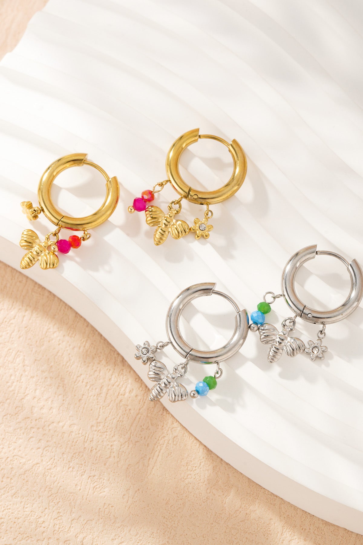 Close-up of gold and silver charm hoop earrings with bee and flower charms and coloured beads on textured cream surface
