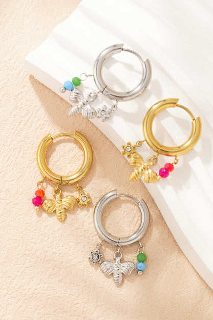 Pair of gold and silver charm hoop earrings with bee, flower charms and colourful beads lying on textured cream surface